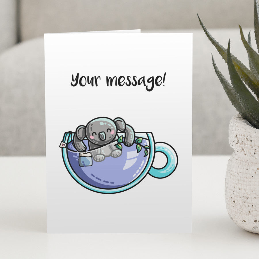 A white greeting card standing on a white table, with a design of a glass teacup of lilac liquid with a koala bear and tea bag in it, and a personalised message above