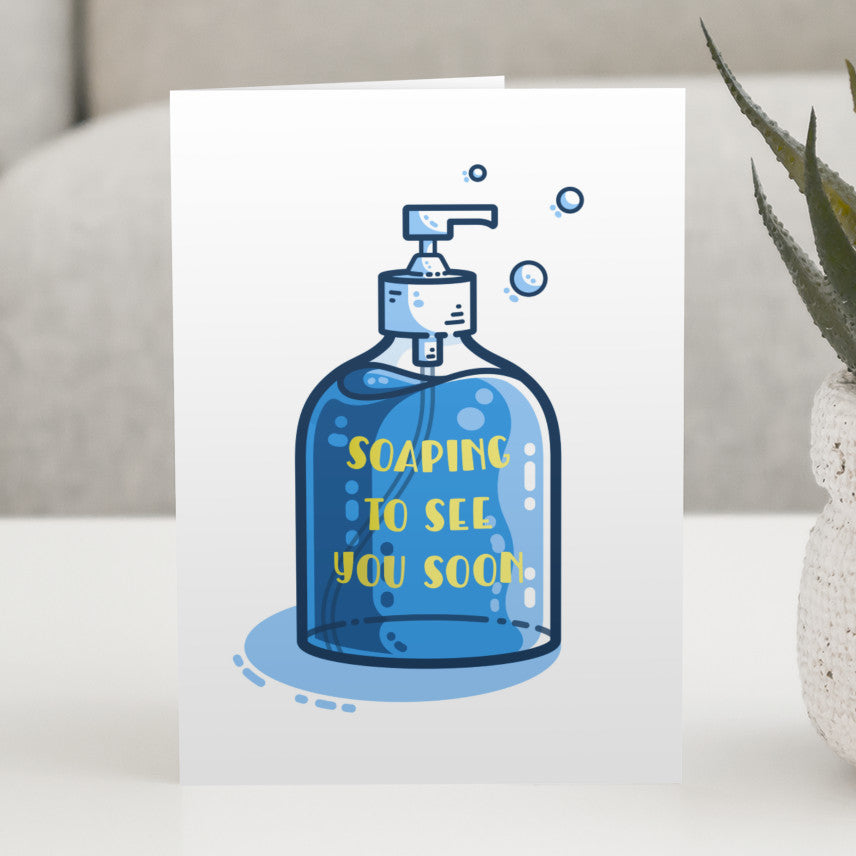A white card standing on a table. The card has a picture of a bottle of blue liquid soap on it and the yellow words on the bottle say soaping to see you soon.
