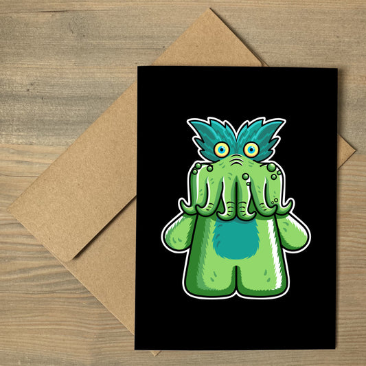 A black greeting card lying flat on a brown envelope, with a design of the StarKid tickle-me-wiggly green plush toy