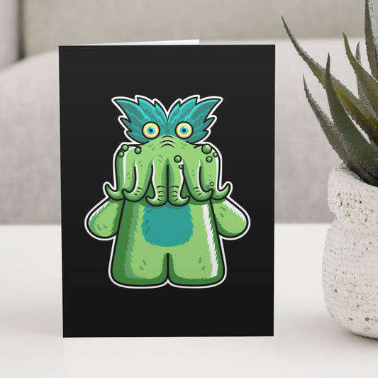 A black greeting card standing on a white table, with a design of the StarKid tickle-me-wiggly green plush toy