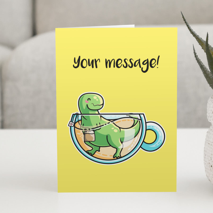 A yellow greeting card standing on a white table, with a design of a cute green tyrannosaurus rex dinosaur holding a teabag in a glass teacup of yellow liquid with a personalised message above