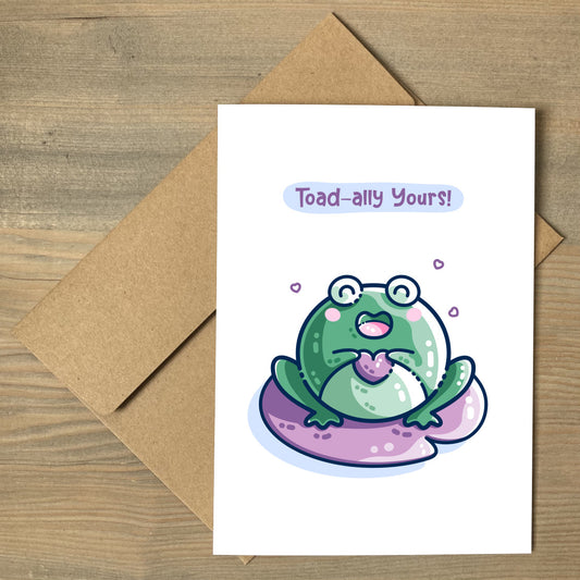 A white greeting card lying flat on a brown envelope and featuring a kawaii cute green toad holding a purple heart and sitting on a purple lilly pad. The words toad-ally yours appear above the toad.