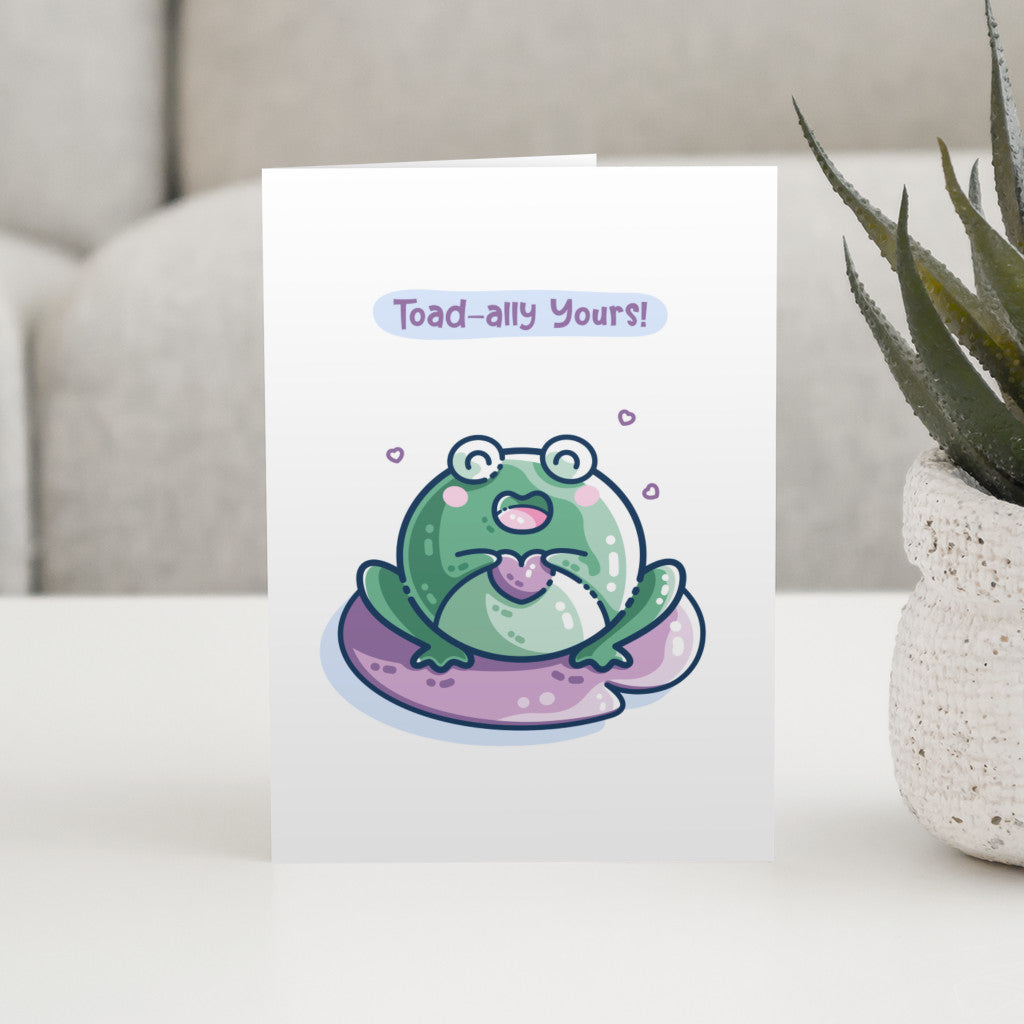 A white greeting card standing on a table and featuring a kawaii cute green toad holding a purple heart and sitting on a purple lilly pad. The words toad-ally yours appear above the toad.