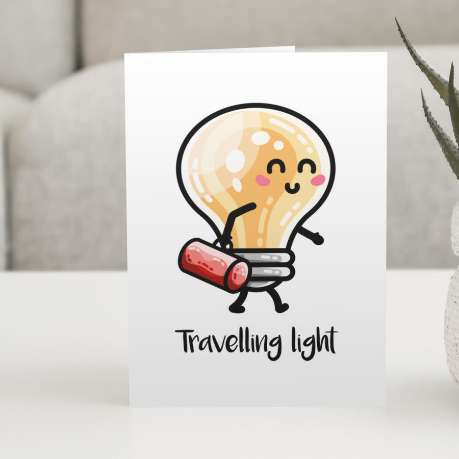 A white greeting card standing on a white table, with a design of a kawaii cute lightbulb walking and carrying a bag with words above saying 'travelling light'