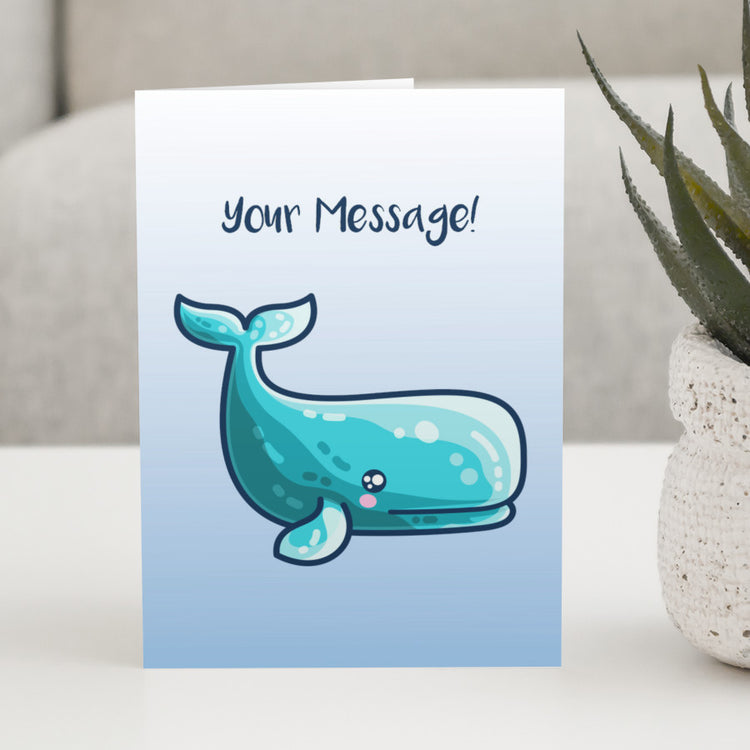 A white to blue gradiant greeting card standing on a white table, with a design of a kawaii cute sperm whale looking to the right and a personalised message above