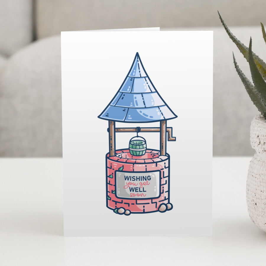 A white greeting card standing on a white table, with a design of a wishing well with blue pointy roof, green bucket, red stone and a sign with the words wishing you get well soon
