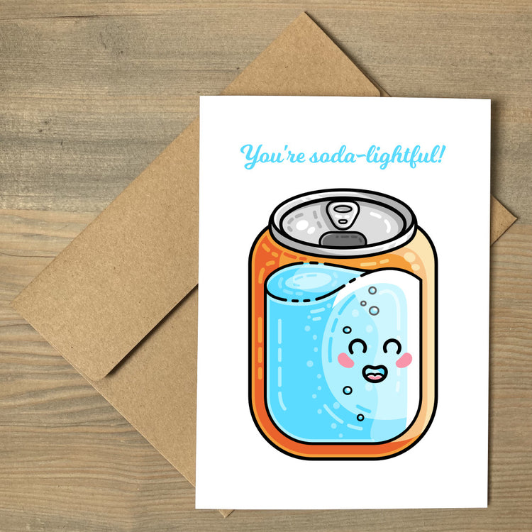 A white greeting card lying flat on a brown envelope, with a design of a kawaii cute blue and orange can of soda with the pun wording above that reads You're soda-lightful!