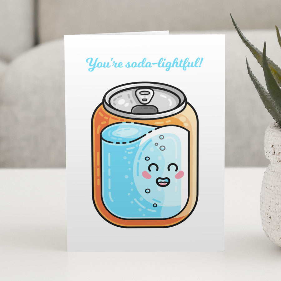A white greeting card standing on a white table, with a design of a kawaii cute blue and orange can of soda with the pun wording above that reads You're soda-lightful!