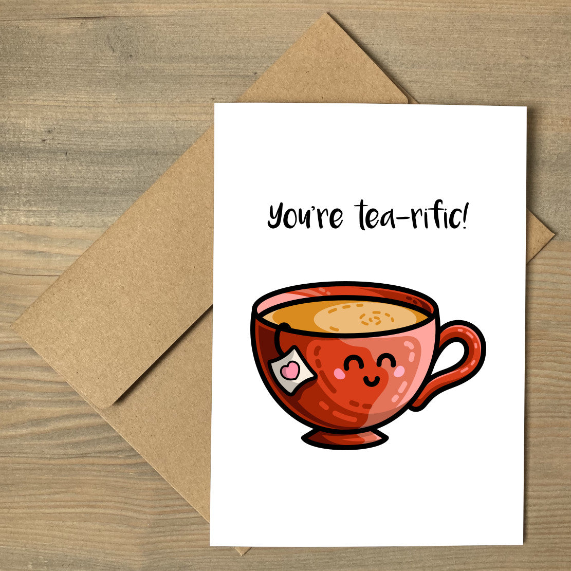 A white greeting card lying flat on a brown envelope, with a design of a kawaii cute red teacup and the wording above reading You're tea-rific!