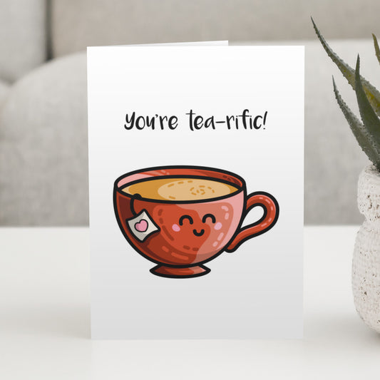 A white greeting card standing on a white table, with a design of a kawaii cute red teacup and the wording above reading You're tea-rific!
